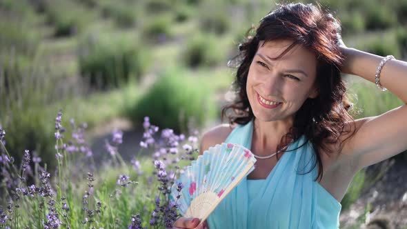 Attractive Adult Woman with Hand Fan in Blue Dress Looking Away Smiling Standing on Lavender Field