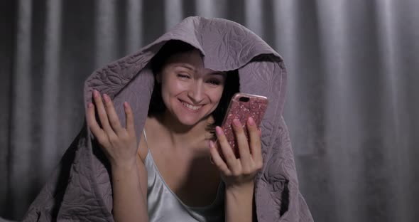 Woman Sitting on Bed Under Blanket. Enjoying Video Chat To Friend on Smartphone