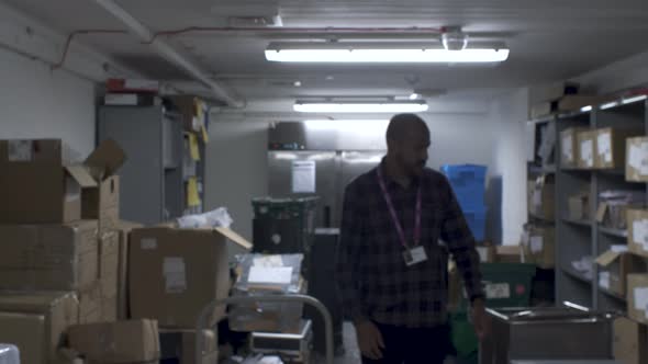 Bald Indian Male Walking Towards Camera In Office Basement And Stopping To Scan Shelves For Item. Ra