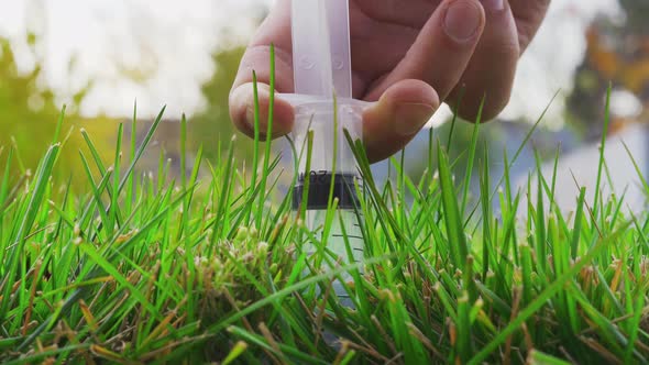 A Hand Inserts a Syringe with a Clear Liquid Into a Bright Saturated Lawn and Squeezes the Contents