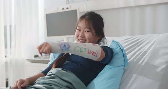 Smiling Asian Preteen Girl Lying in Hospital Bed with Broken Arm in Colored Cast