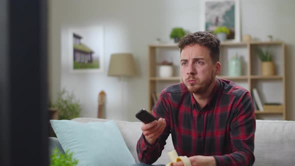 Young Handsome Man Eats Banana and Watching Tv at Home in the Living Room