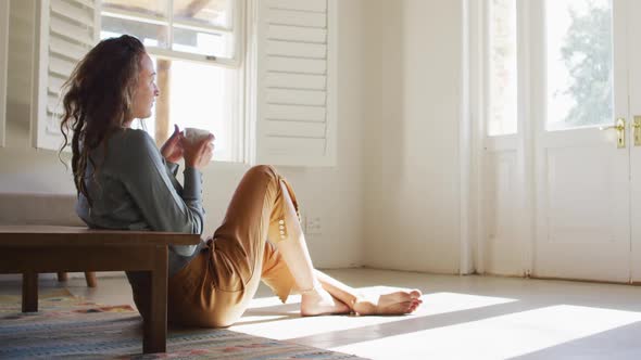 Thoughtful caucasian woman sitting on floor in sunny cottage drinking coffee, looking out of window