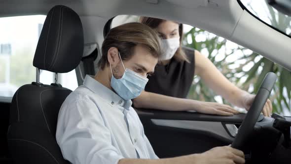 Young Man in Covid-19 Face Mask Sitting on Driver's Seat in Car As Blurred Woman Talking To Buyer in