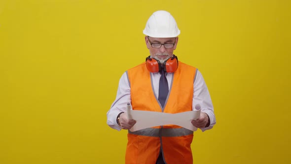 Man Engineer in a Helmet and Vest Holds a Sheet of Drawings in Hands and Speaks Standing on Yellow