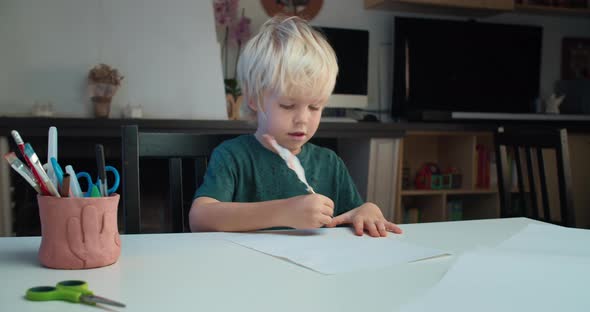 Cute Child Boy Sitting at the Table and Drawing Using Quill Pen in Living Room