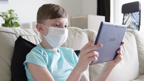 A Young Boy in a Face Mask Works on a Tablet As He Sits on a Couch at Home - Closeup