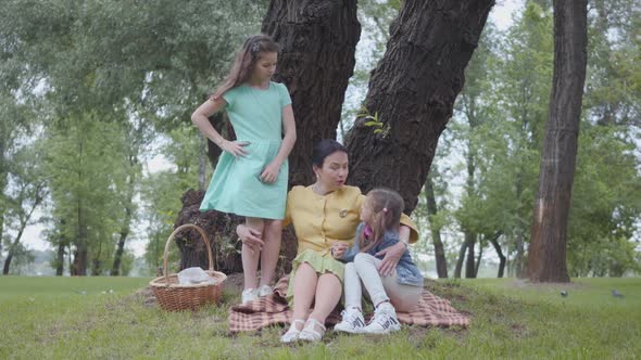 Elegant Senior Woman Sitting on the Blanket Under the Tree in the Park Talking with Granddaughters