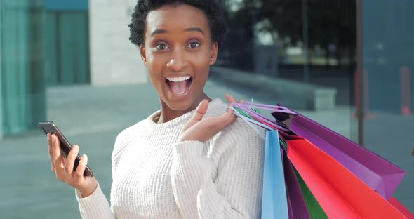 Enthusiastic Woman with Purchases While Shopping Holds Bags with Gifts in Hands Looks at Mobile