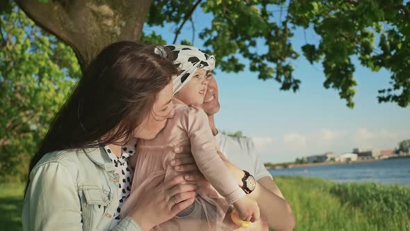 A Young Family Together with a Little Daughter in a Park Under a Tree By the Lake