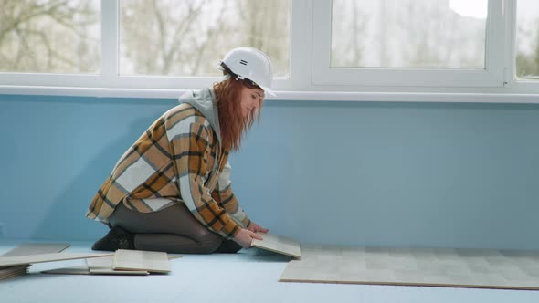 Female Construction Professional in Safety Helmet Lays Laminate Flooring Indoors During Renovation