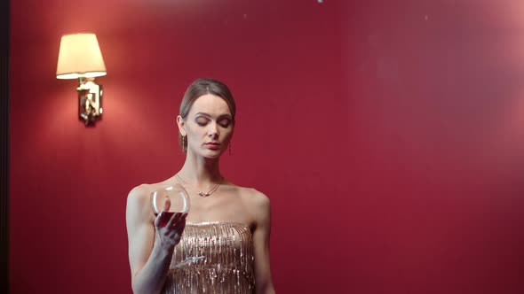 Adorable Female in Shining Dress Posing with Bocal of Wine at Red Room