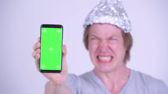 Face of Stressed Young Man with Tinfoil Hat Showing Phone and Looking Scared