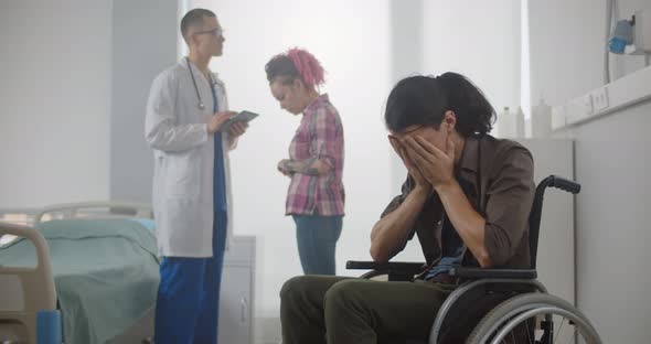 Portrait of Sad Male Patient Sitting on Wheelchair in Hospital While His Wife Consulting Doctor
