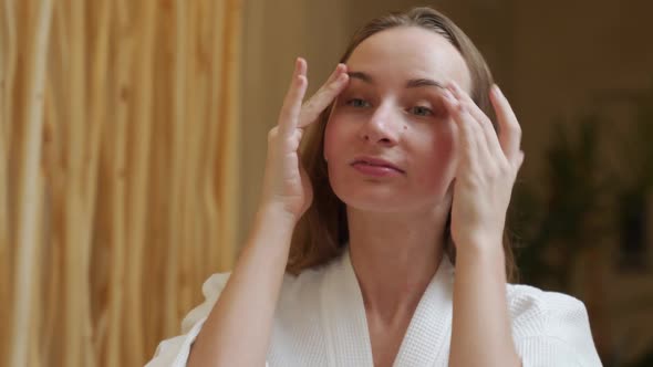 Smiling Beautiful Woman in a Bathrobe Puts a Skin Care Cream on Her Face and Looks in the Bathroom