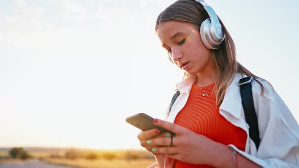 Young Girl Stands on Country Road in Field and Uses Her Phone to Switch Songs in Her Headphones