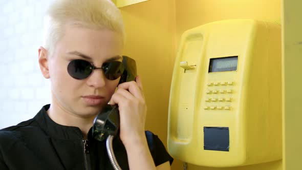 Blonde Woman with Short Haircut Picks Up Phone in Yellow Telephone Booth and Talks on Phone