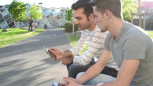 two young multi ethnic men outdoor sitting using smart phone having conversation