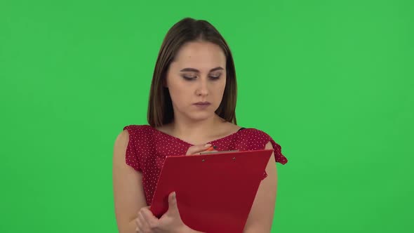 Portrait of Tender Girl in Red Dress Fills Papers in Red Folder with Pensil. Green Screen