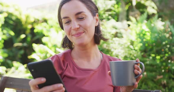 Smiling caucasian woman using smartphone and holding mug of tea sitting in sunny garden