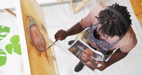 Overhead view of african american male artist painting on canvas looking at photo at art studio