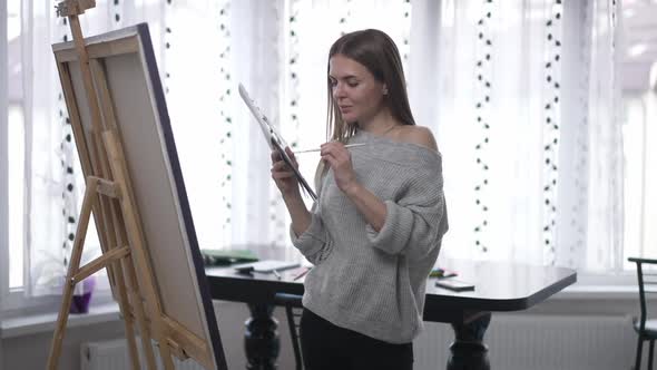 Confident Satisfied Woman Posing with Painting Palette Standing at Easel with Canvas Indoors