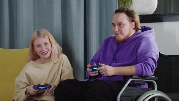 Happy Couple of Blonde Woman and Man in a Wheelchair Playing Game Using Joysticks