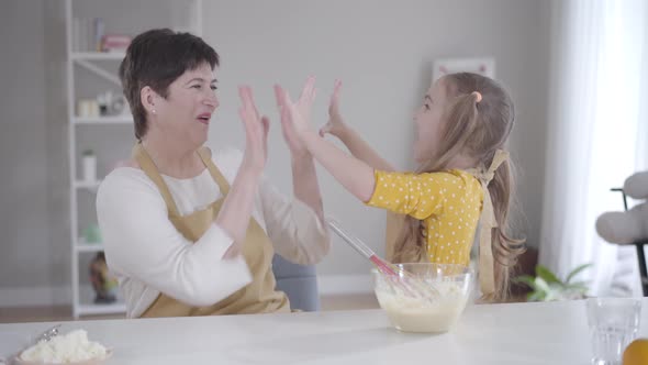 Cute Positive Girl Giving High Five To Grandmother at Kitchen, Happy Woman Hugging Granddaughter