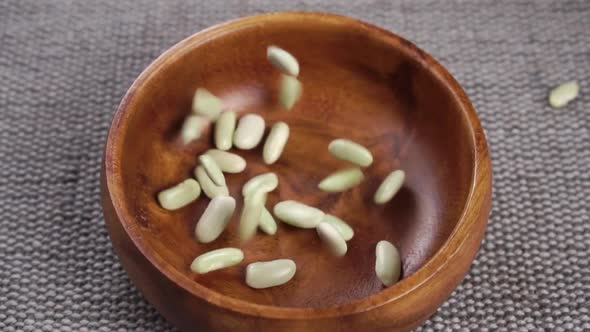 Uncooked green beans fall into a wooden bowl on a gray rustic jute mat close up. Raw legumes