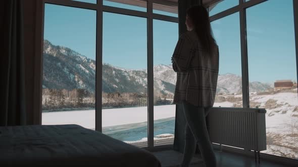 A Woman Goes to the Window in the Bedroom and Drinks Tea From a Mug