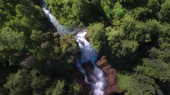 AERIAL shot of sunny turqoise water flowing and falling from lush green forest waterfall