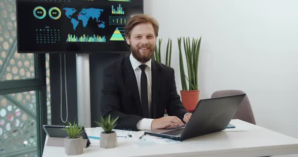 Bearded Ceo in Black Suit Sitting at his Workplace in Office room and Looking Into Camera