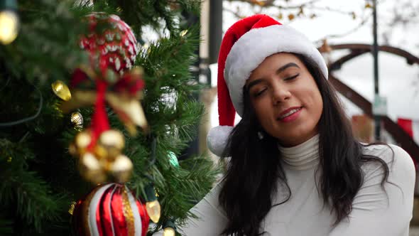 A gorgeous young woman in a Santa Claus hat looking happy and merry next to a holiday Christmas tree