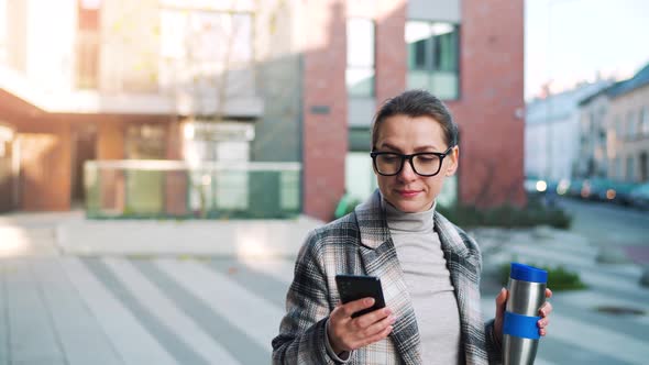 Caucasian Businesswoman with Glasses and in a Coat Walks Through the Business District with Thermo