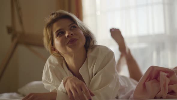 Portrait of Smiling Caucasian Blond Girl Lying on Soft Bed and Shaking Legs. Cheerful Woman in White