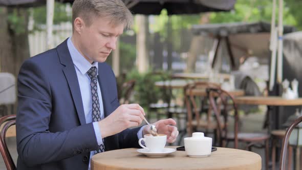 Businessman Mixing Sugar and Drinking Coffee while Sitting in Outdoor Cafe