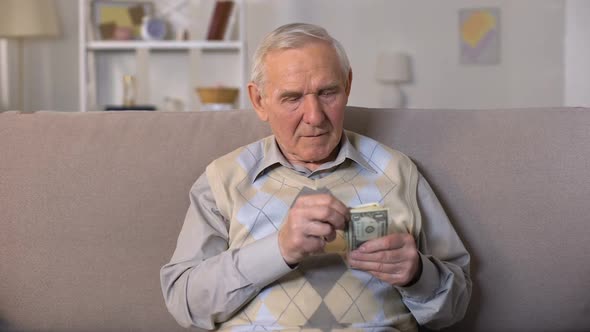 Depressed Old Man Sitting on Sofa, Holding Dollars Banknotes, Social Insecurity