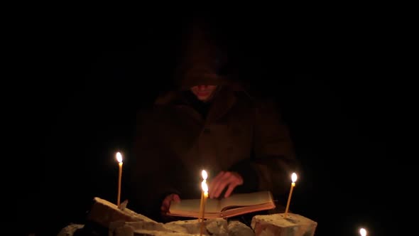 a Man in a Cloak with a Hood is Reading a Hot Book with Candles on the Floor