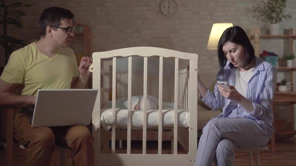 Young Parents Search the Internet and a Newborn in a Baby Bed