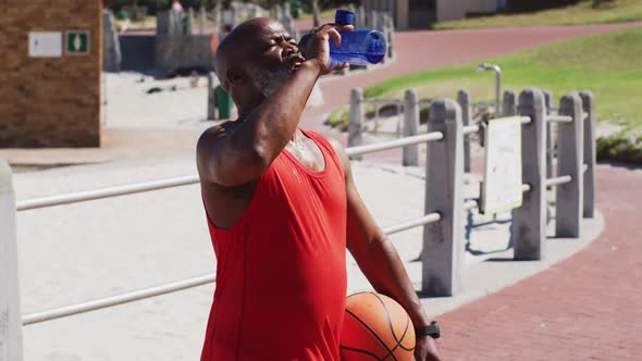 Senior african american man with basketball drinking water on the court near the beach