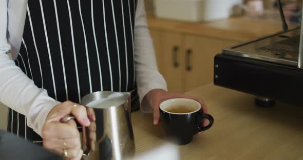 Midsection of caucasian waitress wearing apron, pouring milk into cup of coffee