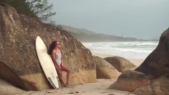 Woman Stands with Surfing Board Leaning Against Rock