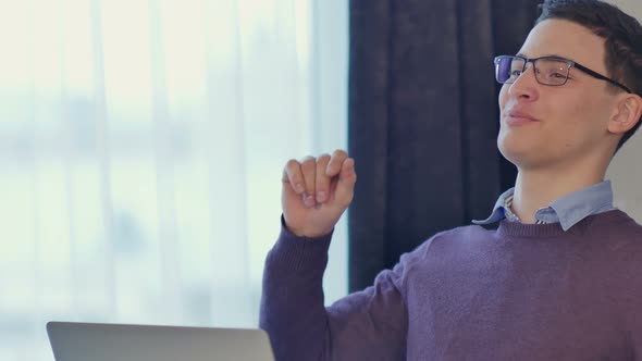 Handsome Young Businessman Sitting at His Desk, Celebrating Success with Arms Raised, Gesturing a