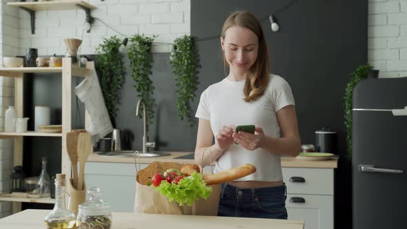 Woman Stands in the Kitchen Near a Paper Bag Full of Fresh Food and Uses a Smartphone App to Deliver