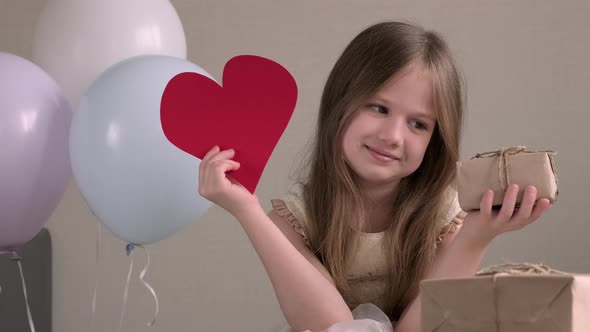 Little Girl Holding Big Red Paper Heart and Zero Waste Gift on Background of Air Balloons