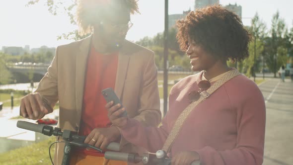 Positive Afro Friends Standing Outdoors with E-Scooters and Using Phone