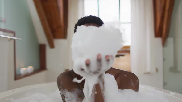 Smiling african american attractive woman taking bath and blowing out foam in bathroom