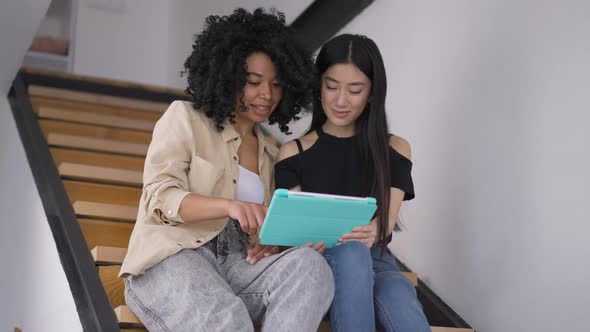 Two Smiling Confident Young Women Surfing Internet on Tablet Talking Sitting in Home Office on