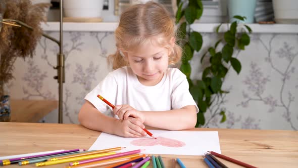 a Little Girl Enthusiastically Draws a Heart with Colored Pencils Sitting at the Table at Home Alone