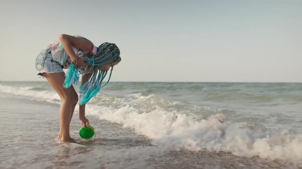 A Girl with African Braids in a Summer Costume Plays on the Beach with Shells Near the Sea with